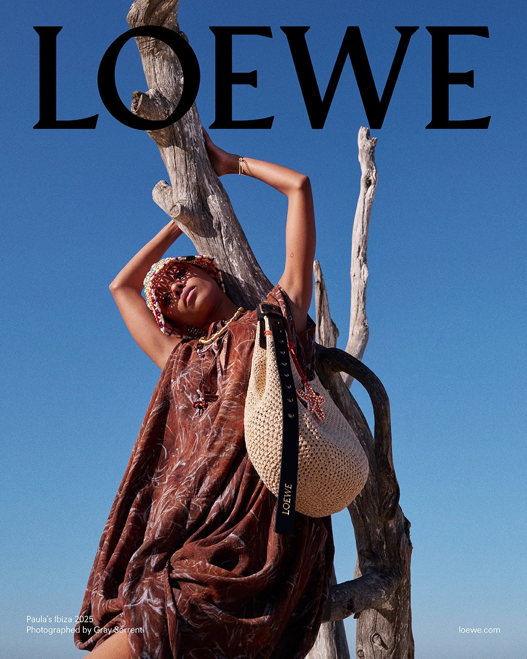 Model in brown patterned dress leaning against a dead tree trunk under a clear blue sky, carrying a straw shoulder bag.