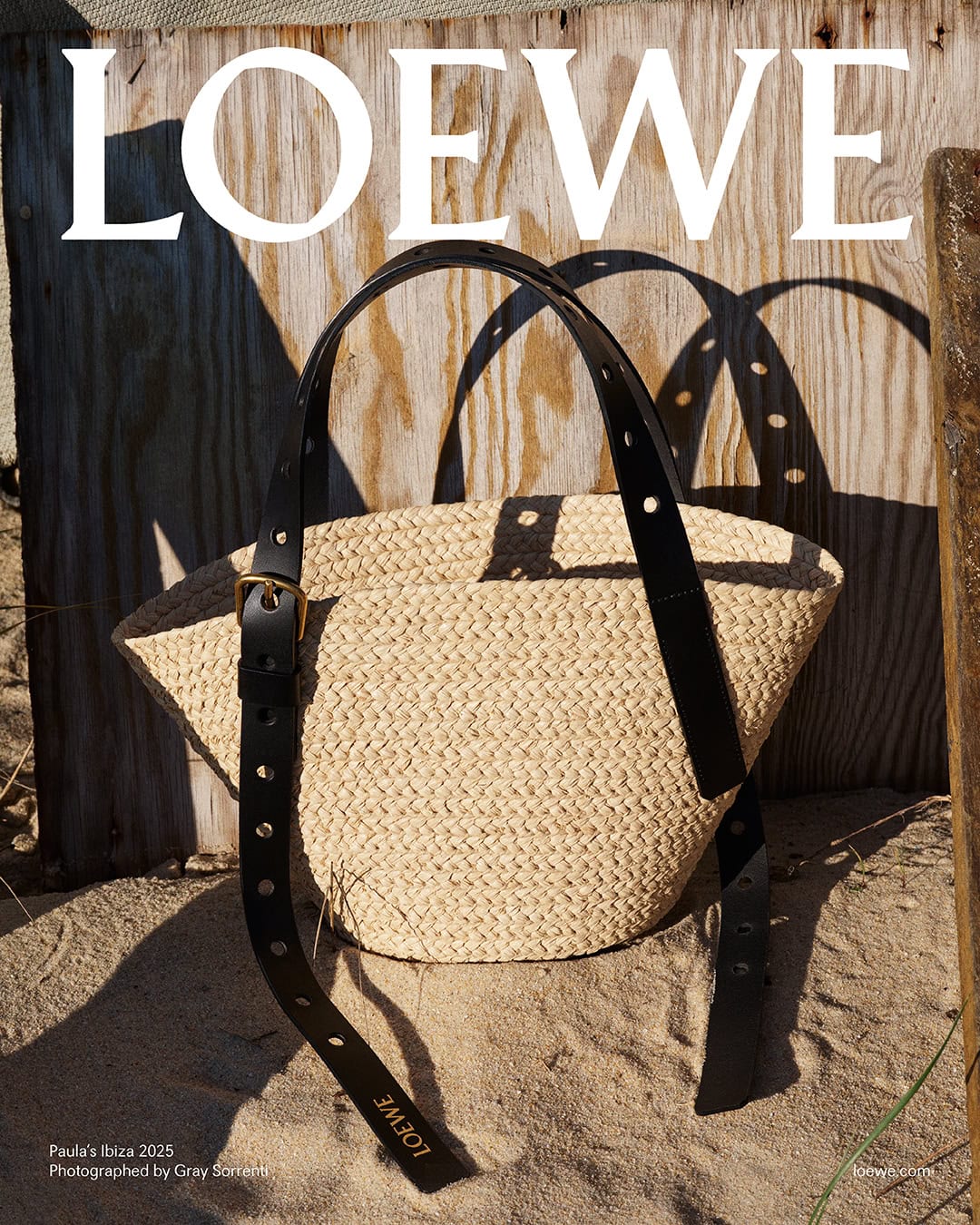 Loewe woven straw basket bag with black leather straps resting on sand against weathered wooden boards.