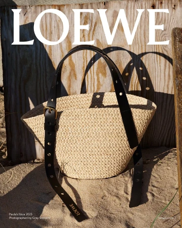 Loewe woven straw basket bag with black leather straps resting on sand against weathered wooden boards.
