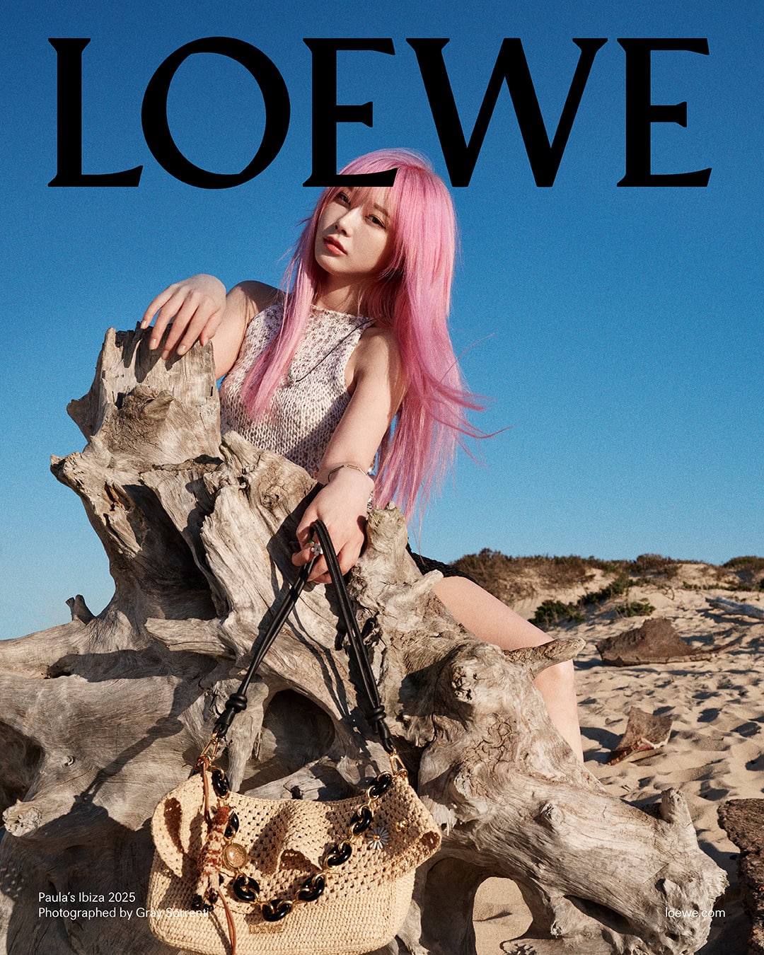 Woman with long pink hair sitting on weathered driftwood on a sandy beach holding a woven straw handbag under a clear sky