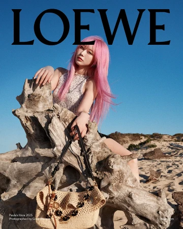 Woman with long pink hair sitting on weathered driftwood on a sandy beach holding a woven straw handbag under a clear sky