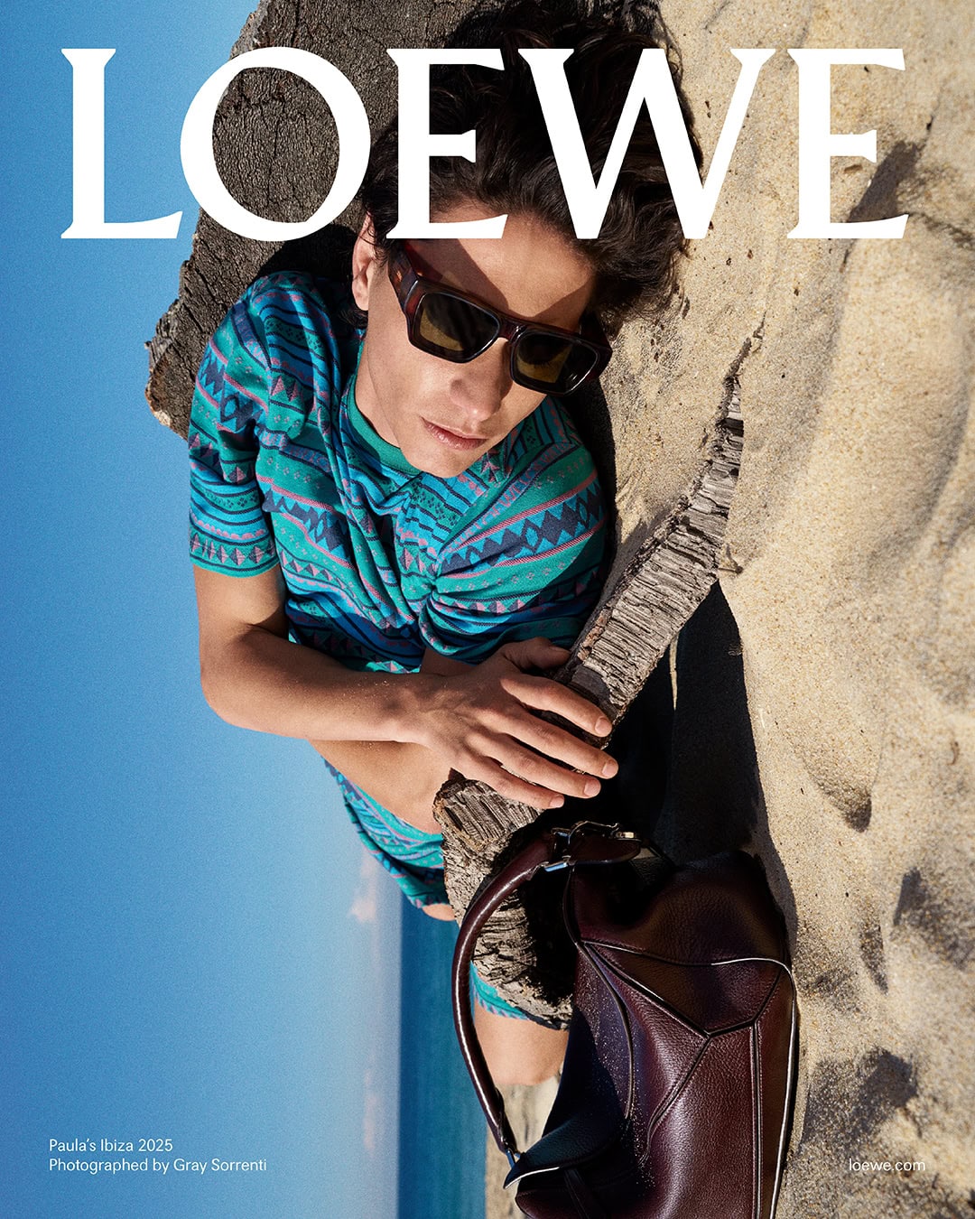 Man wearing sunglasses and a patterned blue t-shirt lying on a sandy beach next to a dark leather handbag.