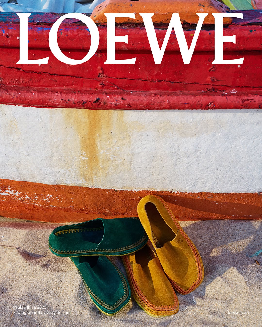 Green and yellow suede loafers resting on sand in front of a red, white, and orange painted wooden boat hull.