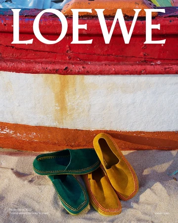 Green and yellow suede loafers resting on sand in front of a red, white, and orange painted wooden boat hull.