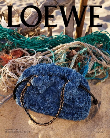 Textured blue handbag resting on sandy beach with driftwood and green netting in background under Loewe headline