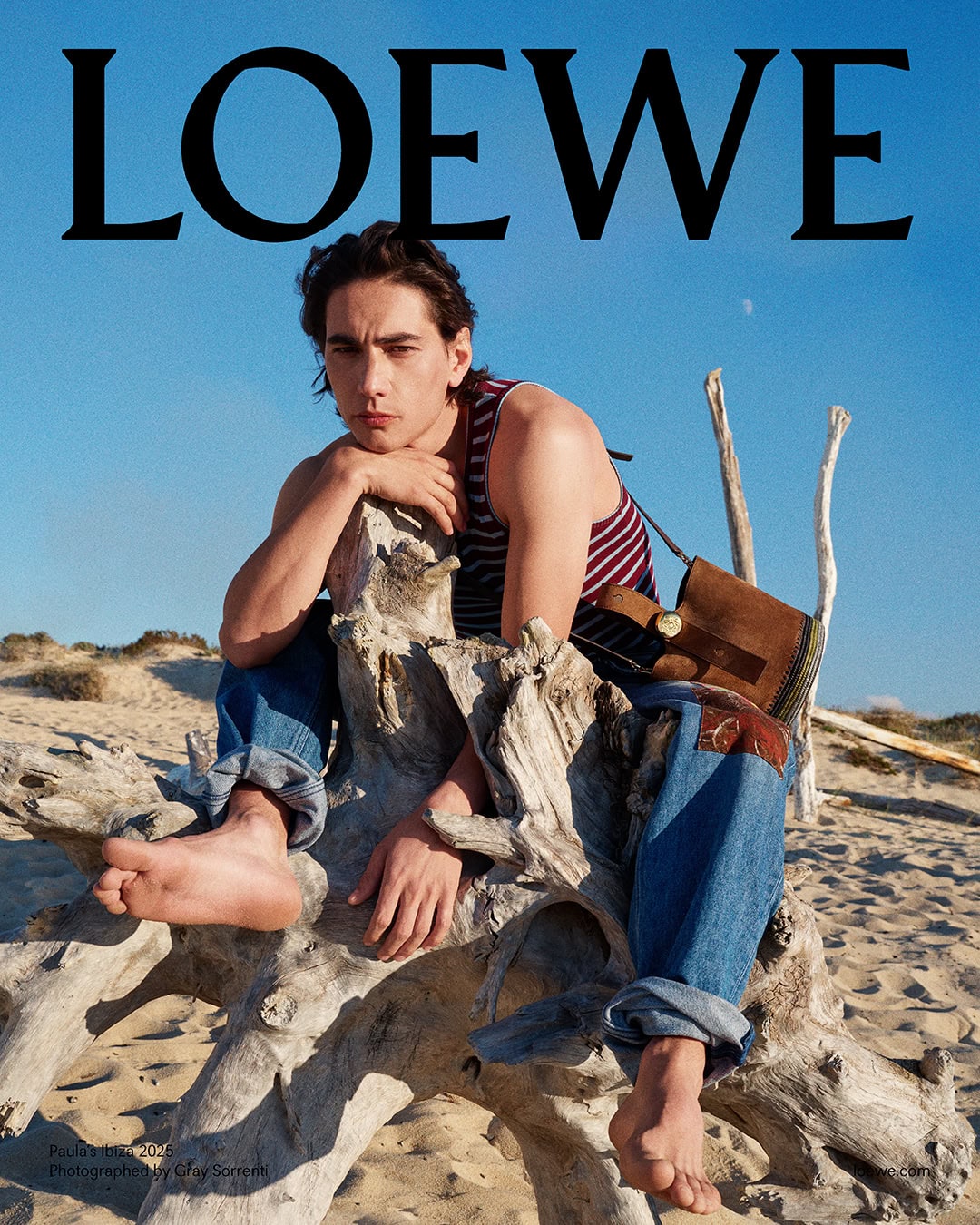 Young man in a striped tank top and denim jeans sitting barefoot on a weathered driftwood log on a sandy beach.