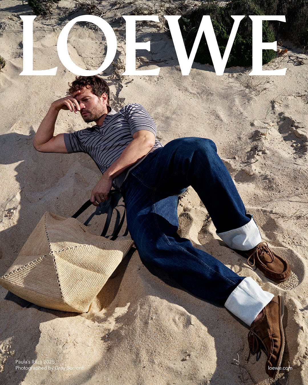 Man in striped polo and cuffed jeans reclining on sand dunes next to a straw tote bag