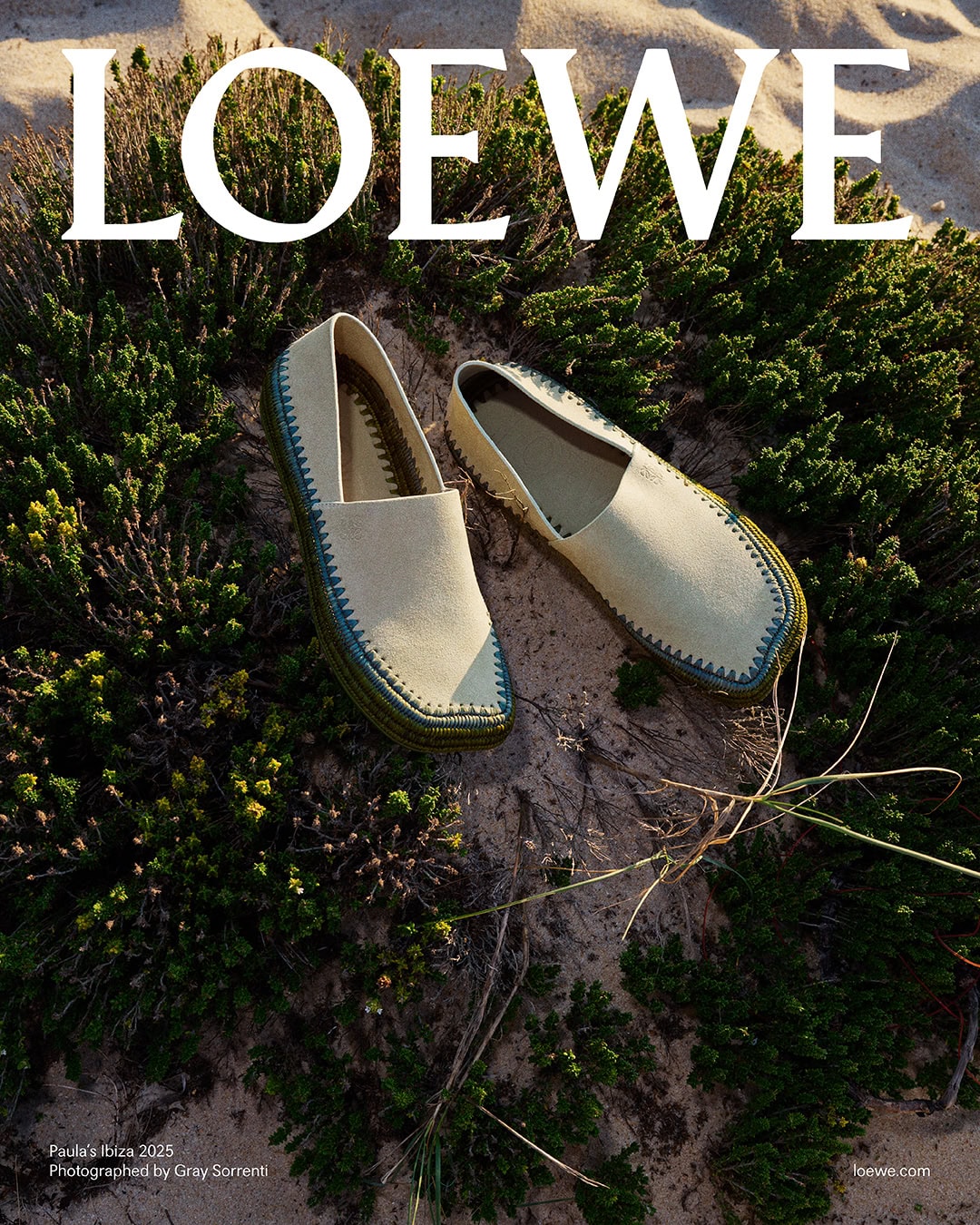 Pair of canvas slip-on espadrilles with decorative stitching resting on sand among coastal shrubs.