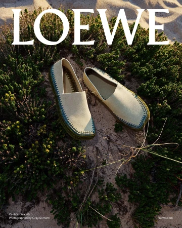 Pair of canvas slip-on espadrilles with decorative stitching resting on sand among coastal shrubs.