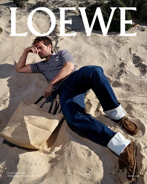 Man in striped polo and cuffed jeans reclining on sand dunes next to a straw tote bag