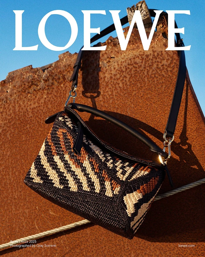 Loewe woven handbag hung against a textured rusty metal surface under a bright blue sky.