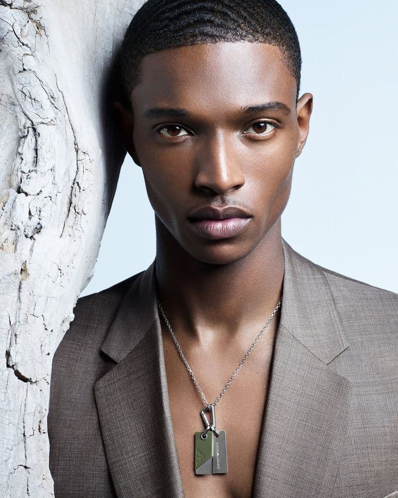 Close-up portrait of a Black male model wearing a brown blazer and dog tag necklace against a pale background