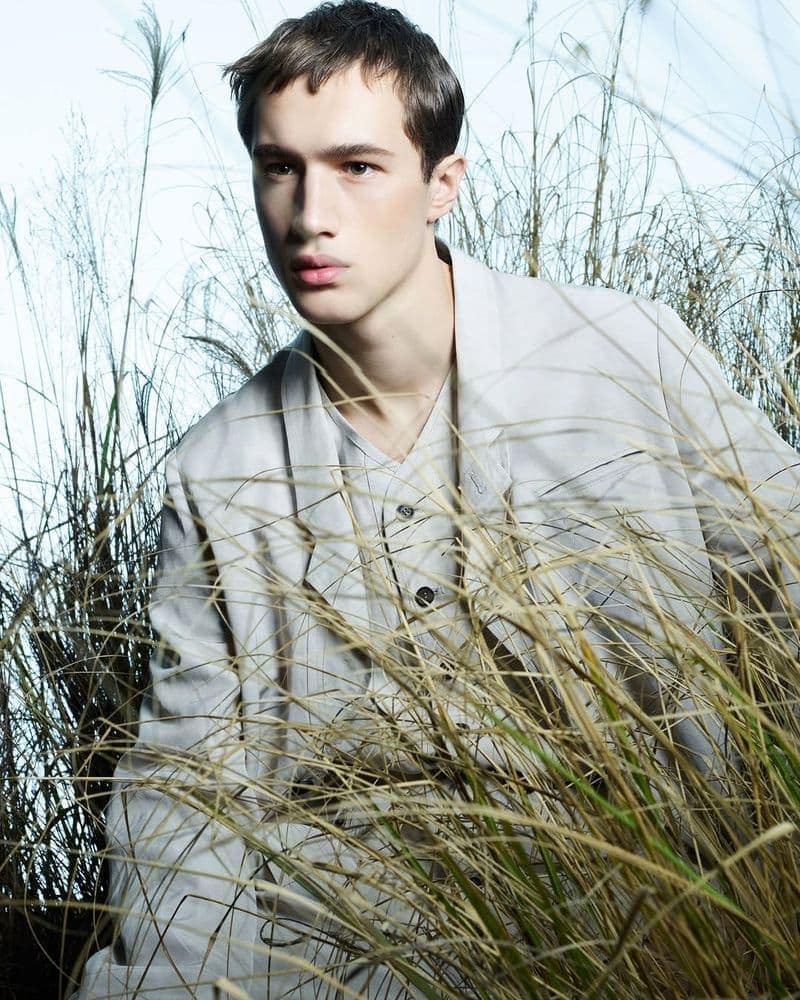 Young man wearing a light grey suit jacket amidst tall dry grass for Emporio Armani spring 2025 ad campaign.