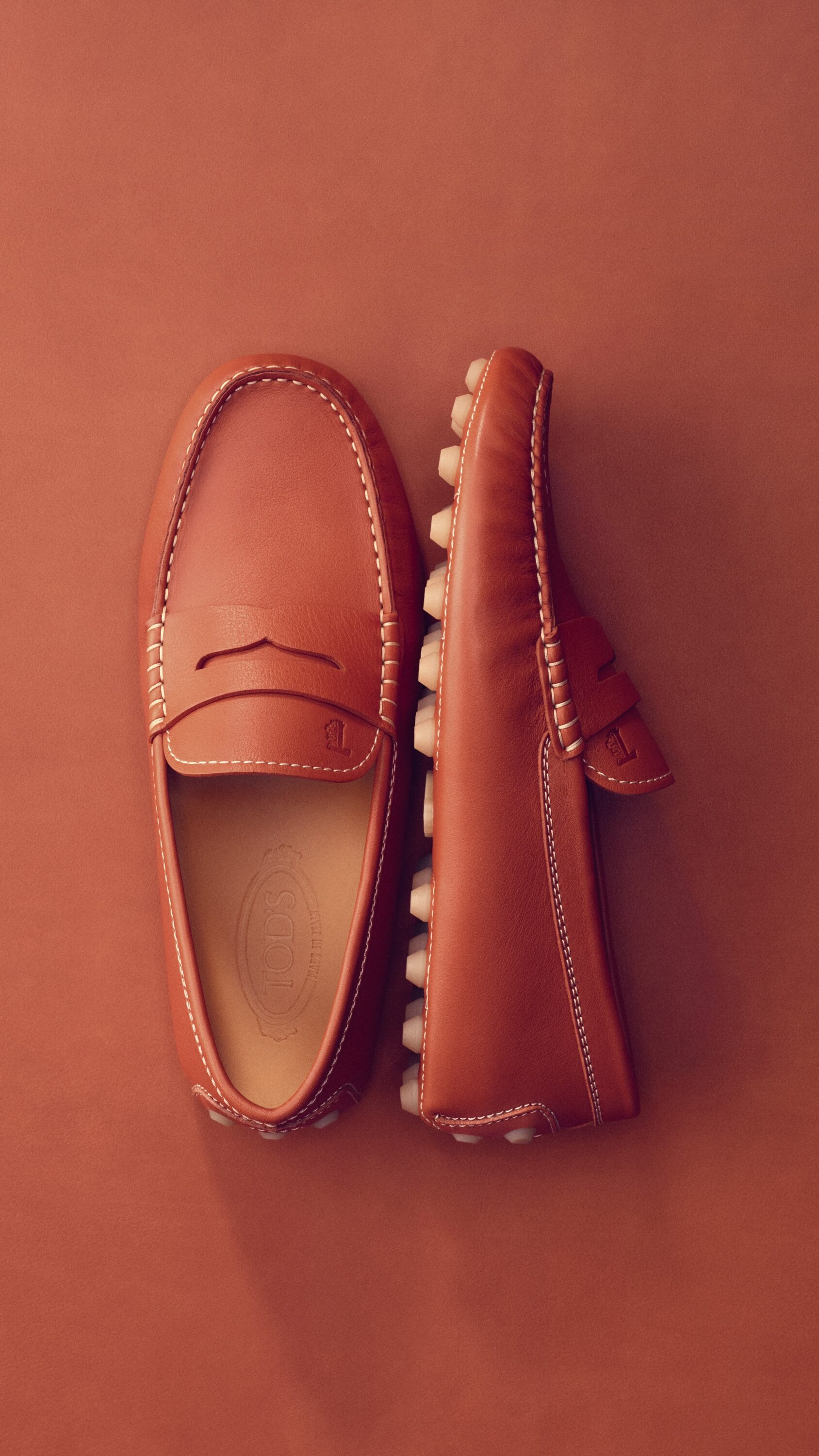 Top-down view of a pair of brown leather loafers with white stitching against a warm terracotta background.