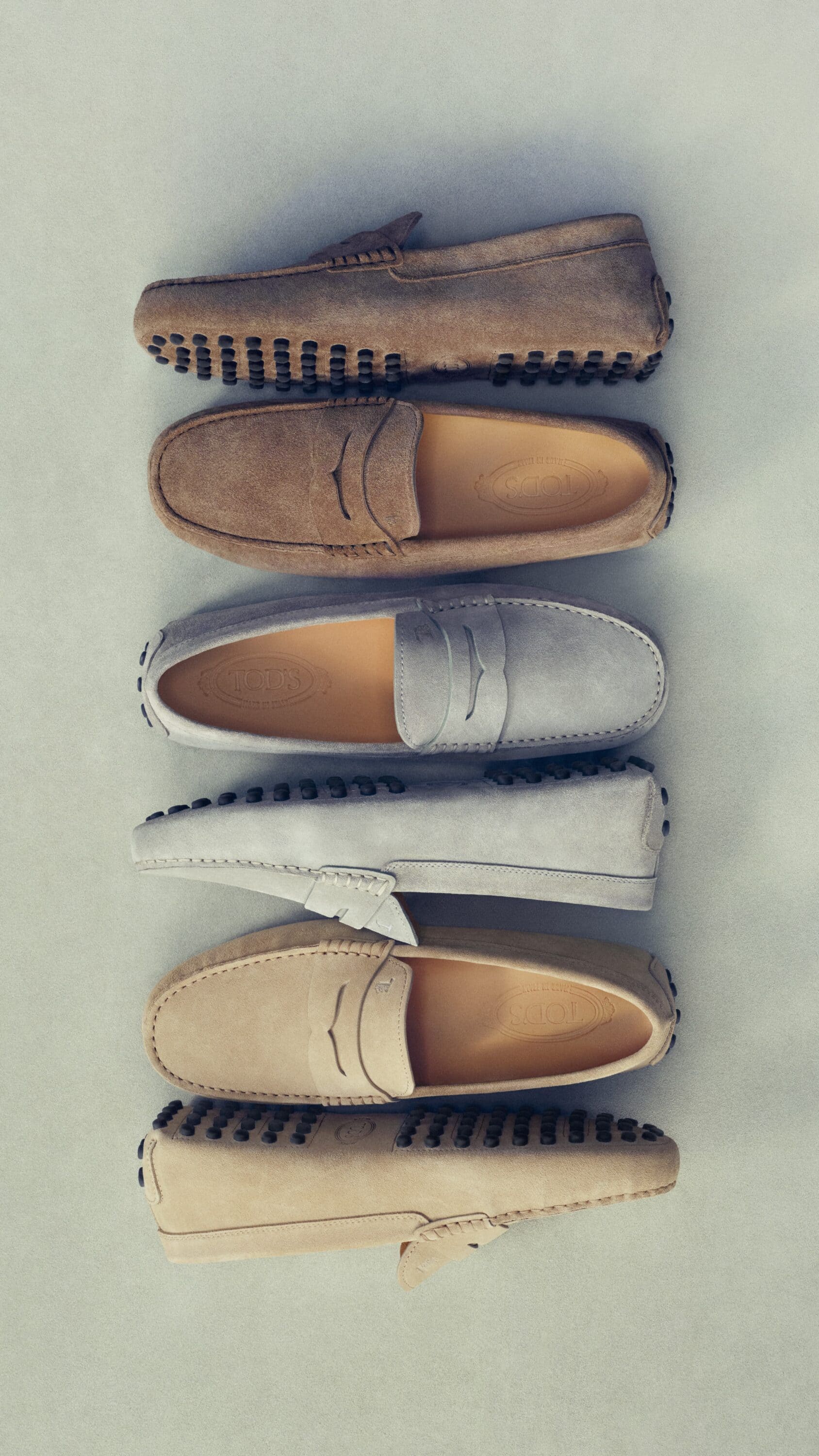 Top-down view of six suede loafers arranged in a vertical line against a neutral surface for Tod's Spring 2025 campaign.