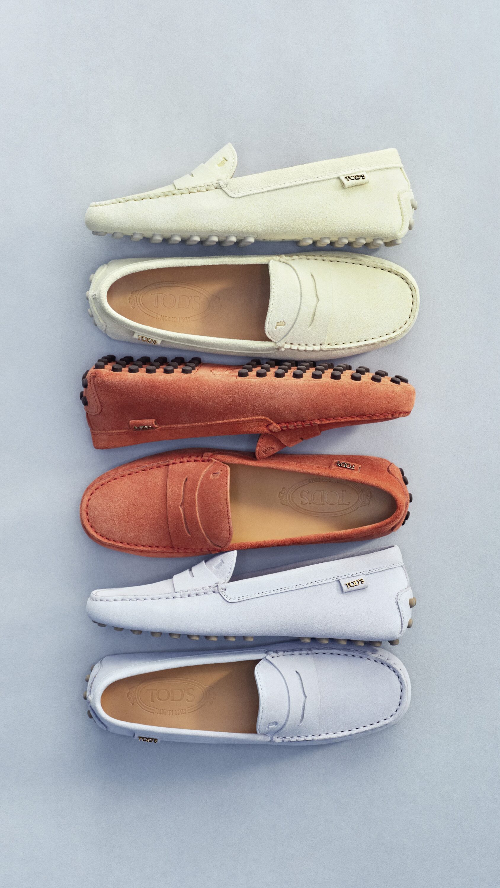 Six pairs of Tod's Gommino driving shoes arranged in a vertical row against a neutral grey seamless background.