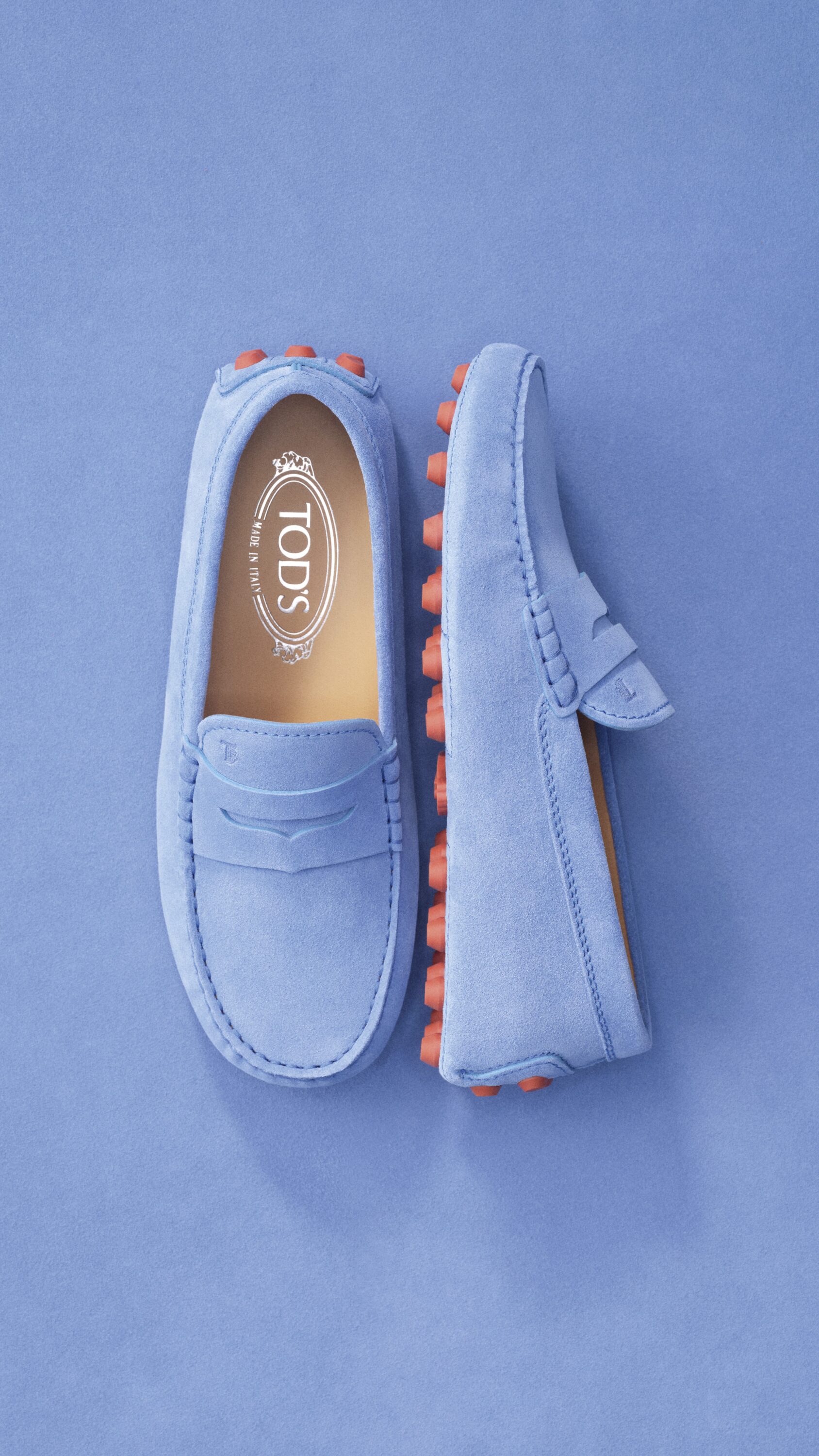 Top-down view of light blue suede Tod's loafers featuring distinctive rubber pebble soles against a blue textured backdrop.