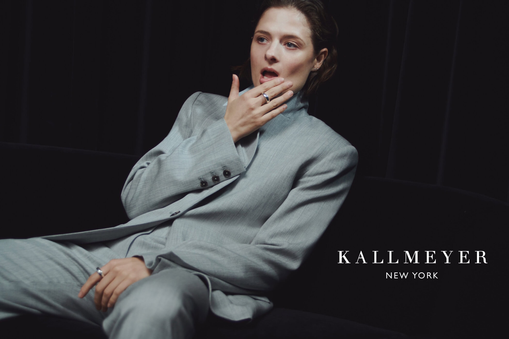 Model wearing a grey suit jacket with hand near mouth against a dark background for Kallmeyer campaign