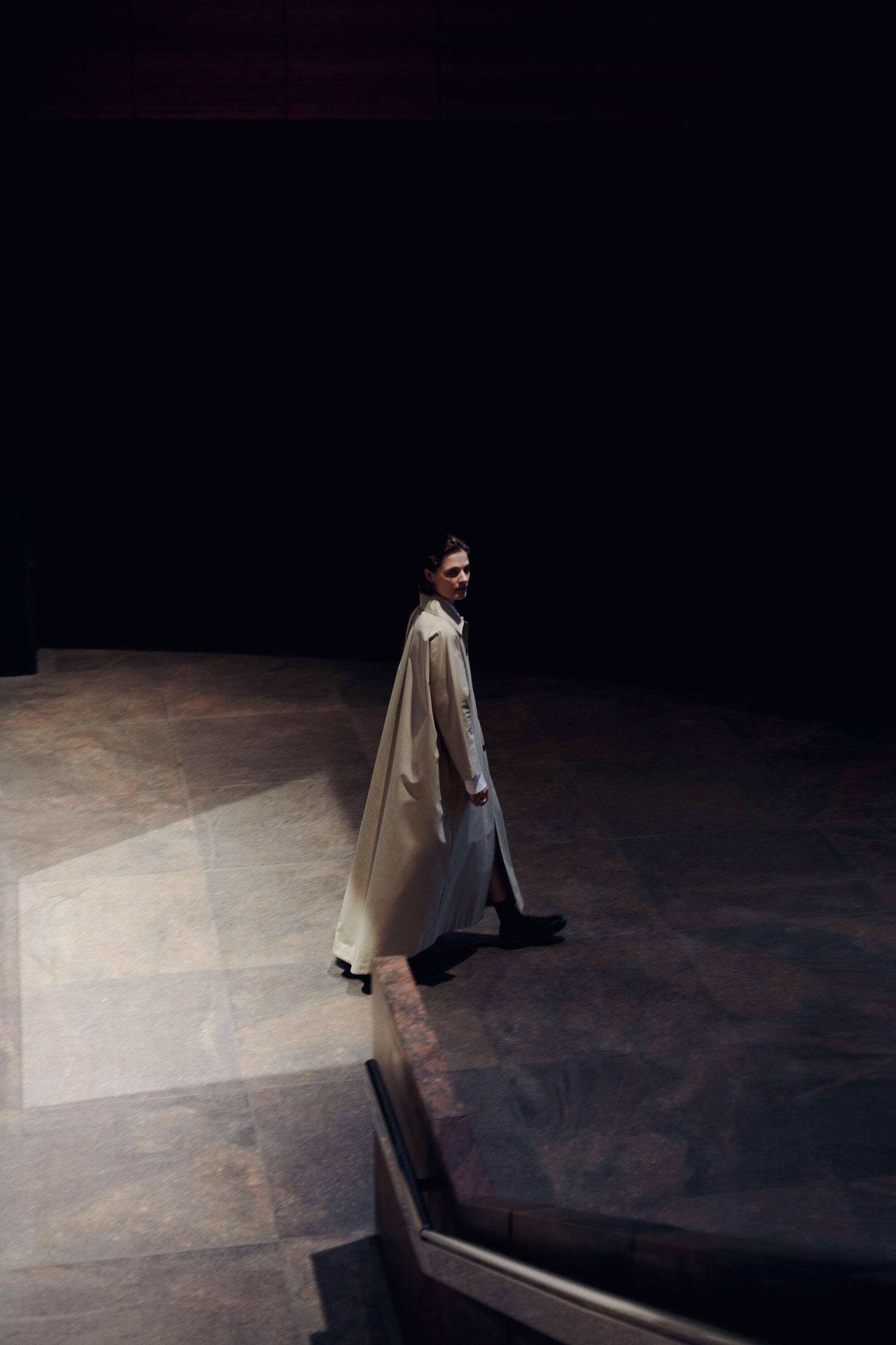 Model in a long white coat walking across a dark tiled interior space for Kallmeyer