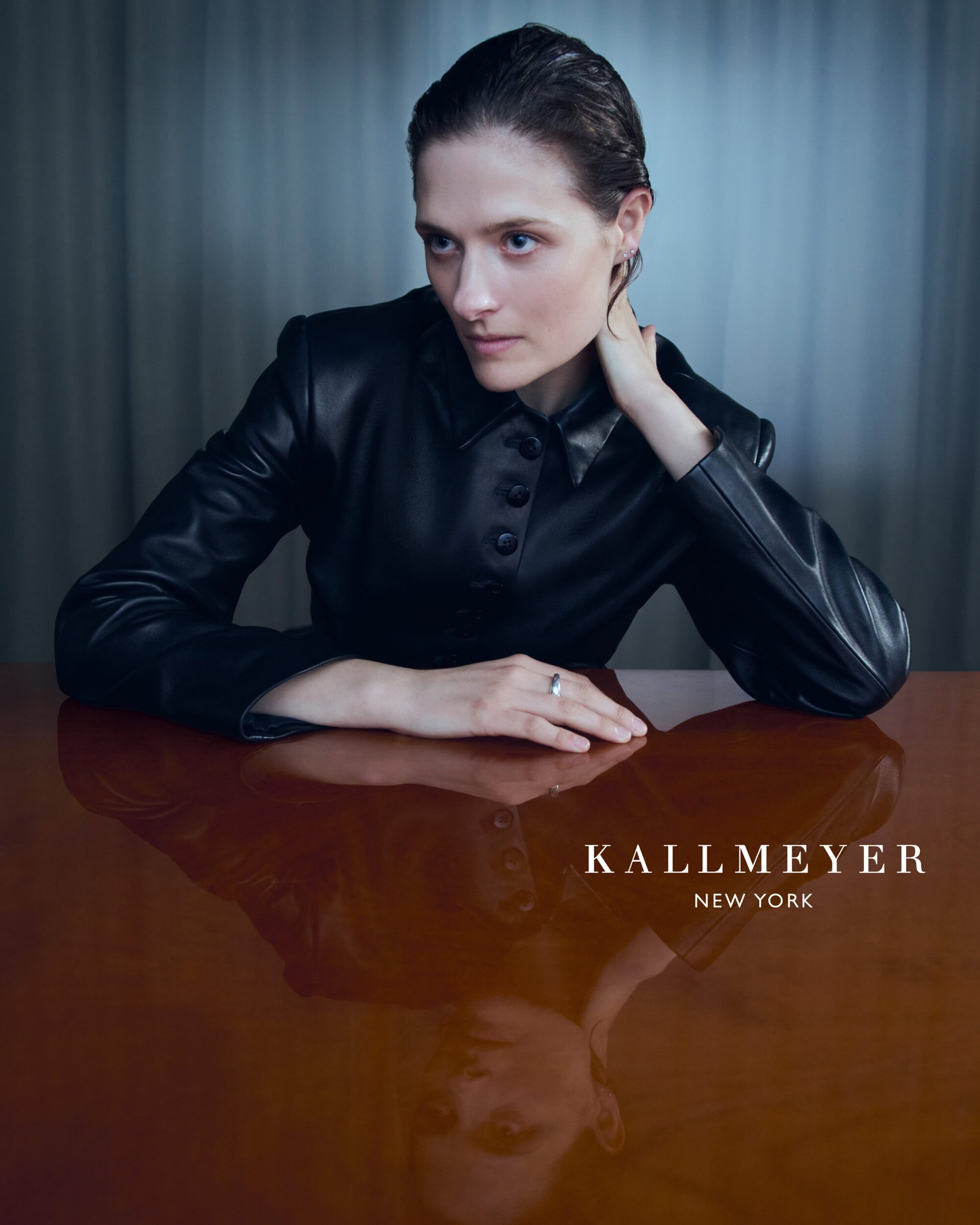 Model in black leather jacket resting arms on a glossy wooden table with a visible reflection, shot for Kallmeyer Spring 2025 campaign.