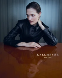 Model in black leather jacket resting arms on a glossy wooden table with a visible reflection, shot for Kallmeyer Spring 2025 campaign.