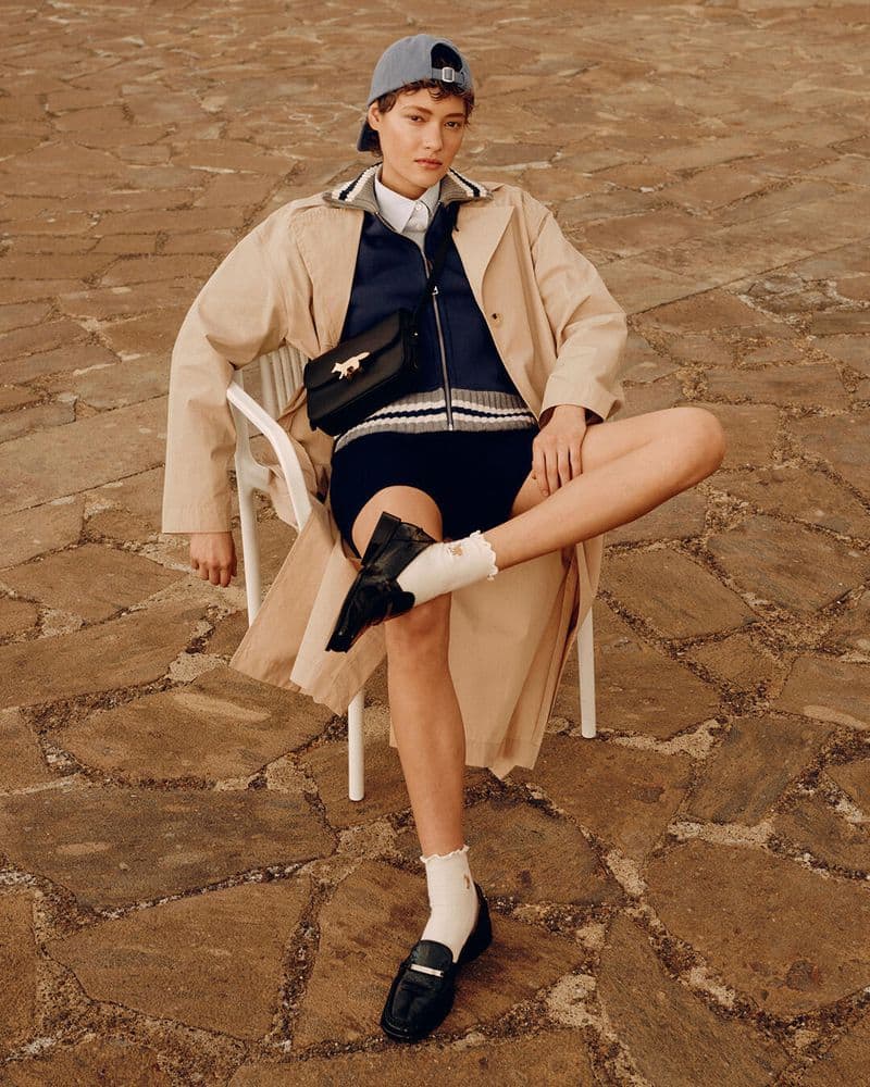 Model seated on a white chair outdoors wearing a beige trench coat, blue cap, dark zip sweater, shorts, and loafers.