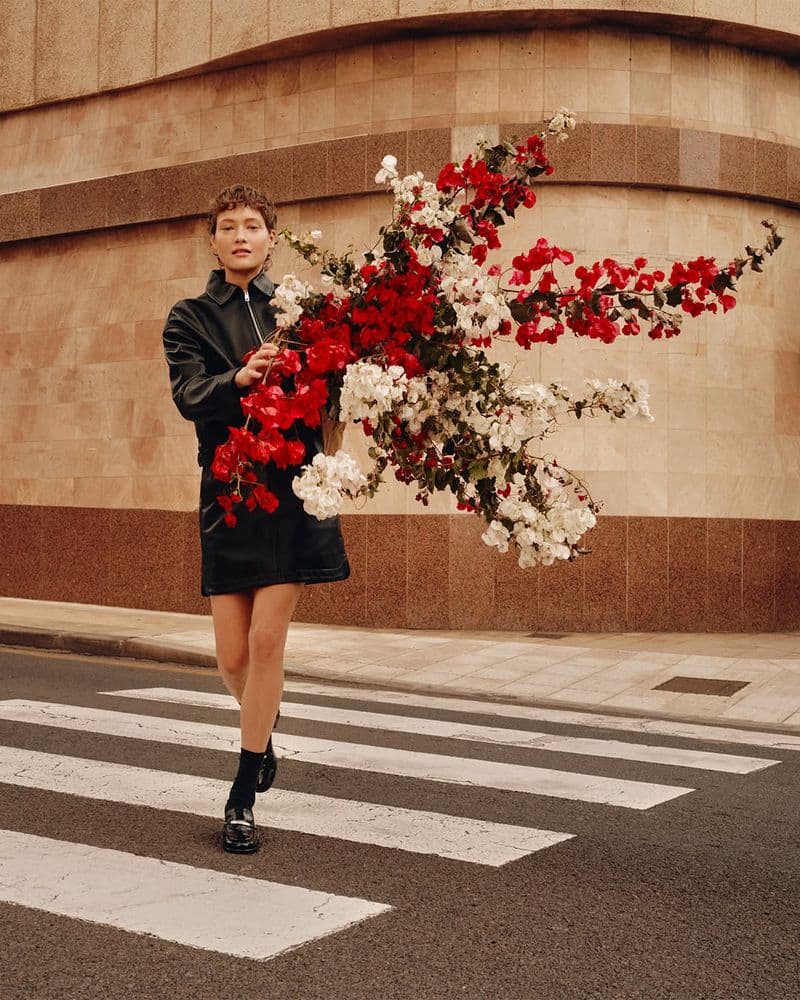 Model wearing a black leather jacket holding a large branch of red and white flowers while crossing a street