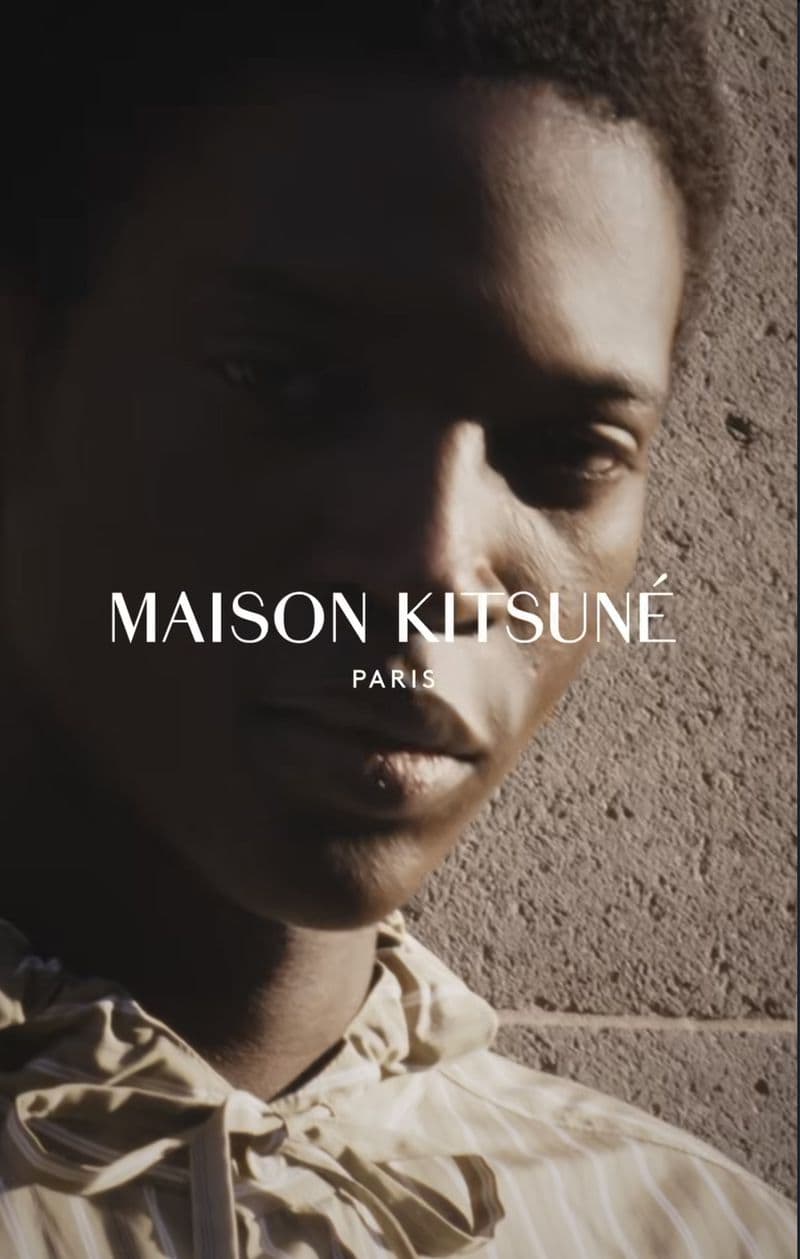 Close-up portrait of a model wearing a striped hooded garment against a rough stone wall for Maison Kitsune Spring 2025 campaign