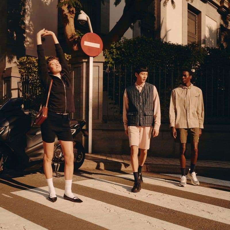 Three models walking across a street crosswalk in sunlight for the Maison Kitsune Spring 2025 campaign.