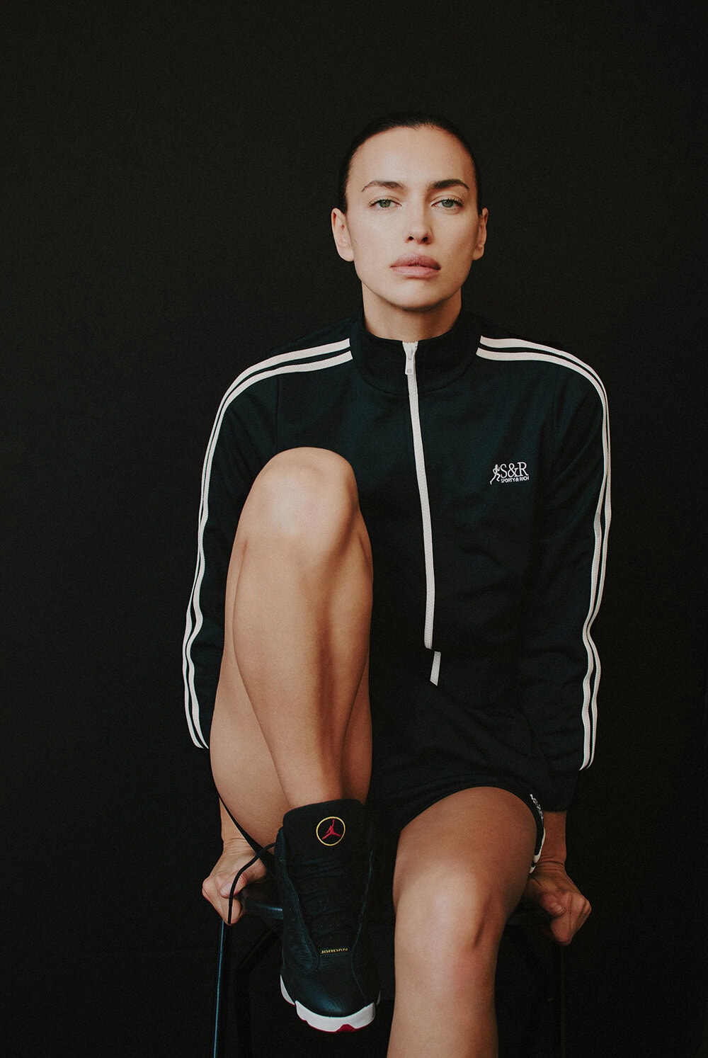 Model Irina Shayk wearing a dark athletic track jacket against a plain dark studio background