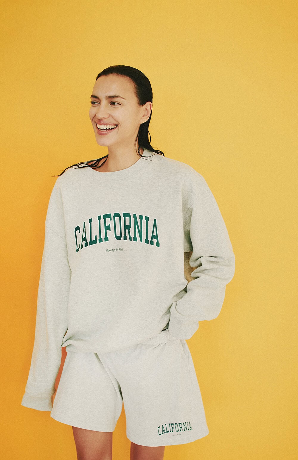 Model Irina Shayk wearing a light grey California sweatshirt against a solid yellow background.