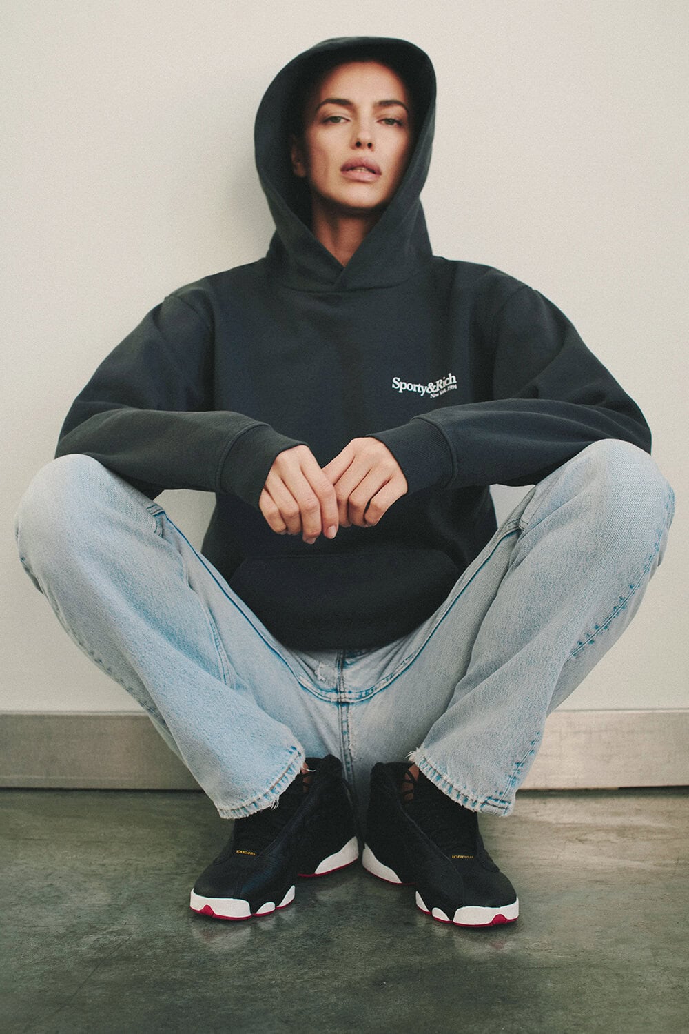 Model Irina Shayk wearing a dark hooded sweatshirt and light wash jeans crouching against a neutral wall