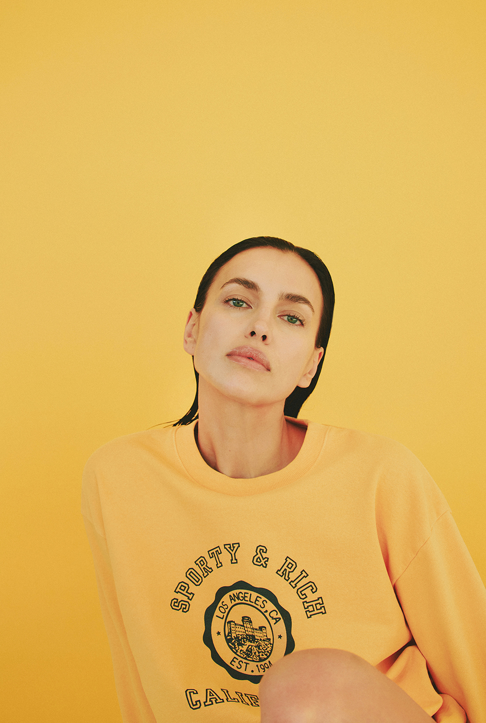 Irina Shayk wearing a yellow sweatshirt with green branding against a warm yellow backdrop