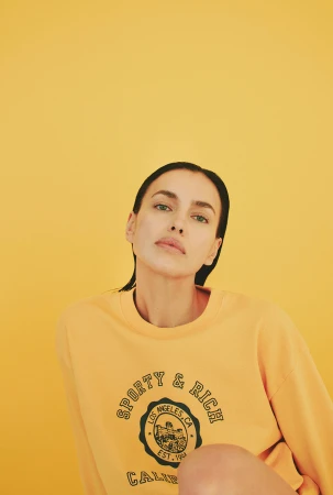 Irina Shayk wearing a yellow sweatshirt with green branding against a warm yellow backdrop