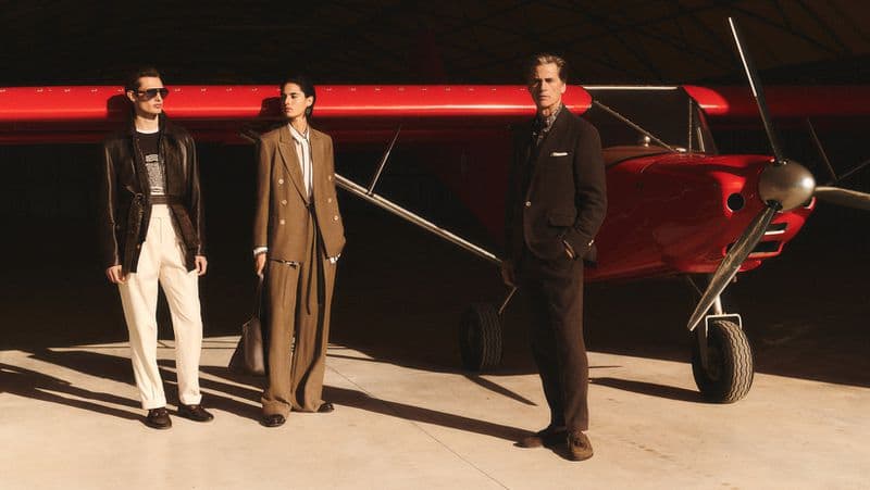 Three models posing in front of a bright red airplane inside a hanger for Brunello Cucinelli spring 2025 ad campaign.
