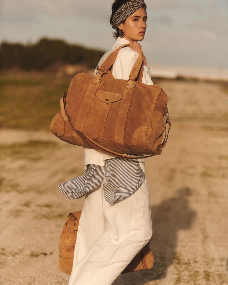 Model carrying a large suede travel bag wearing white trousers and headband outdoor
