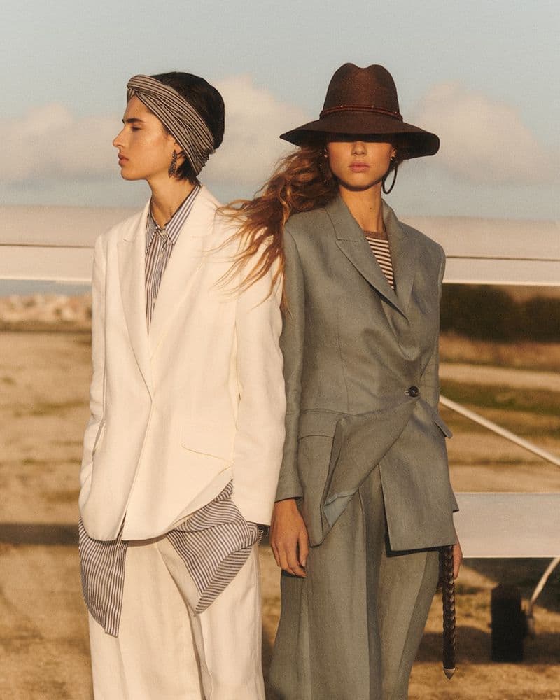 Two female models in tailored blazers standing outdoors with a light aircraft wing in the background.