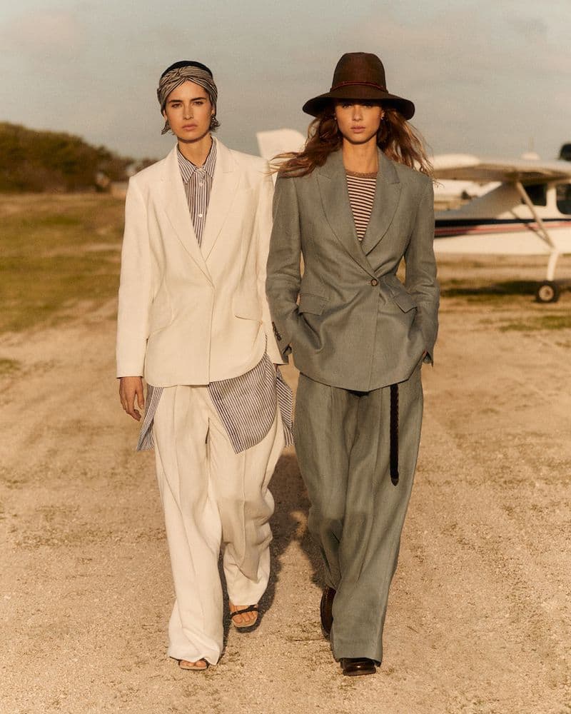 Two models wearing tailored suits walk on a dirt airstrip in front of a small airplane, styled for Brunello Cucinelli.