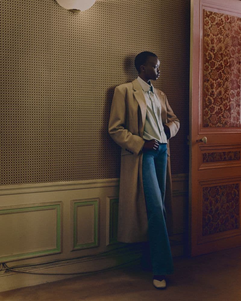 Model leaning against a patterned wall wearing an oversized beige coat and blue trousers