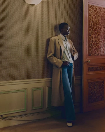 Model leaning against a patterned wall wearing an oversized beige coat and blue trousers