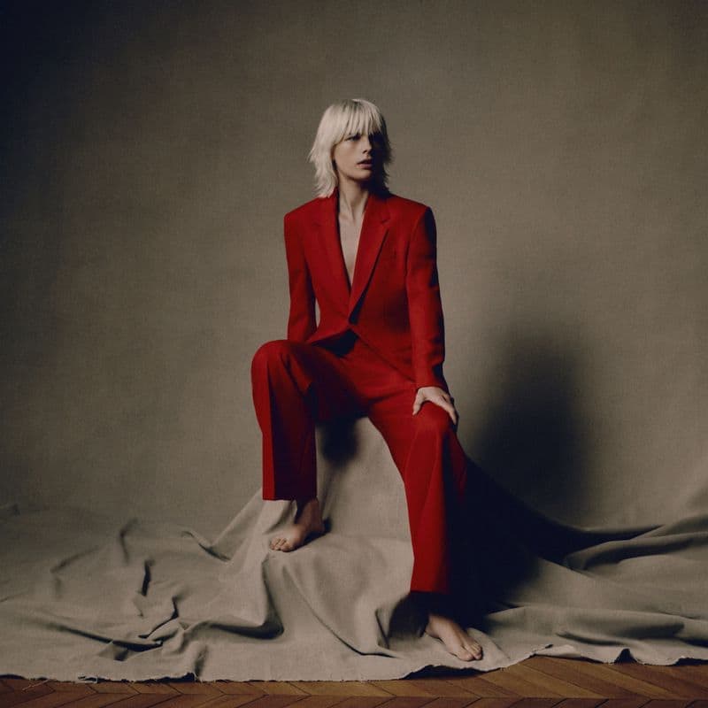 Model with blonde hair wearing a red suit seated on a draped taupe fabric backdrop