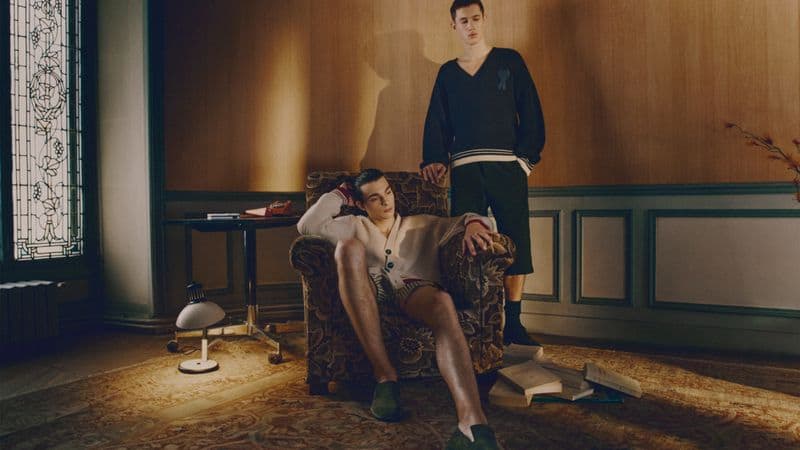 Two men posing in a traditional interior, one seated in an armchair and the other standing beside him