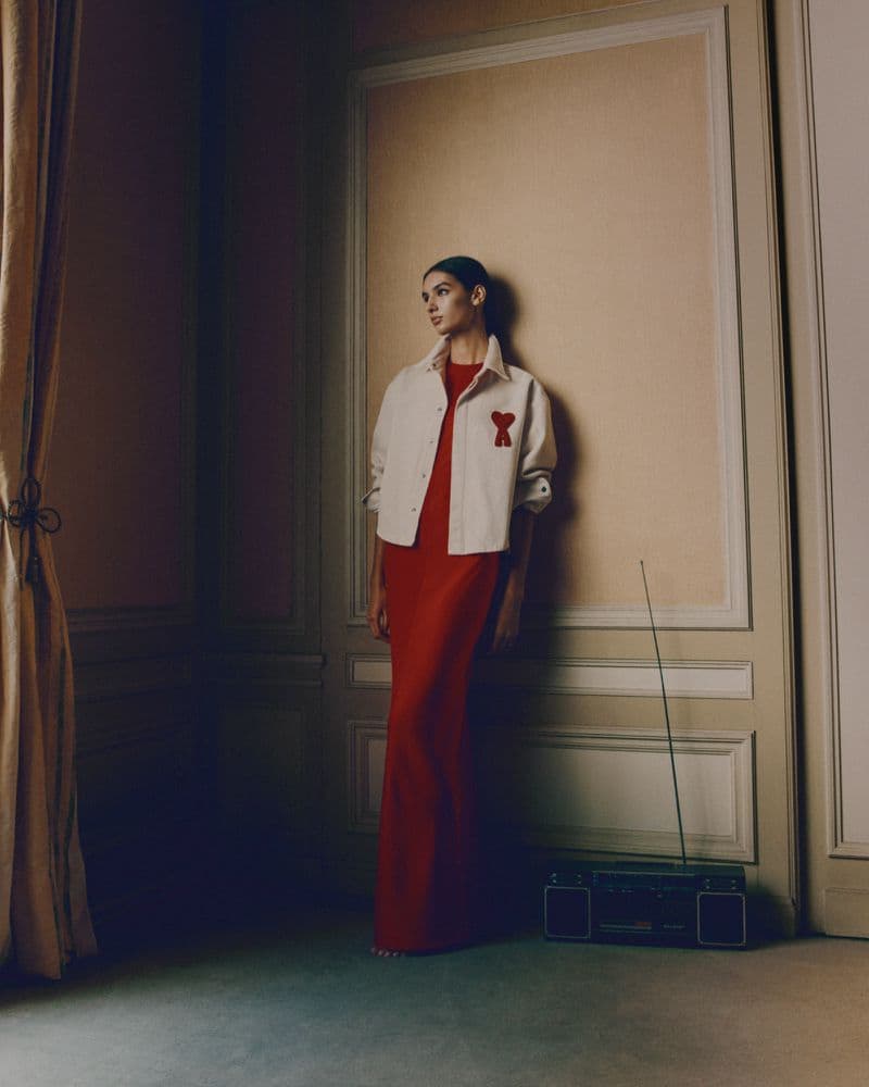 Model wearing an off-white jacket and long red dress leaning against a wall in an interior room with a radio on the floor