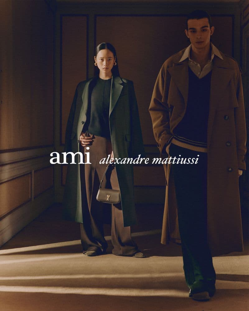 Two models wearing structured coats stand in a dimly lit hallway with panelled walls and warm sunlight casting diagonal beams across the floor.