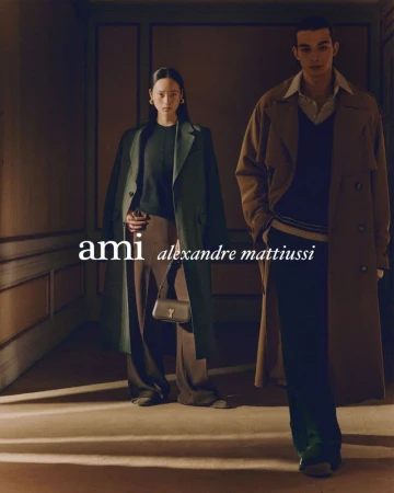 Two models wearing structured coats stand in a dimly lit hallway with panelled walls and warm sunlight casting diagonal beams across the floor.