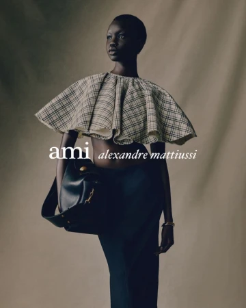 Model wearing a structured plaid cape and black skirt holding a dark leather handbag against a neutral studio backdrop