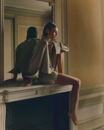 Woman in oversized striped shirt seated on a white mantelpiece with a large mirror reflecting her figure