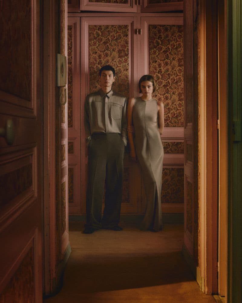 Two models standing in a domestic hallway with pink paneled walls and patterned wallpaper for Ami Paris Spring 2025 campaign.