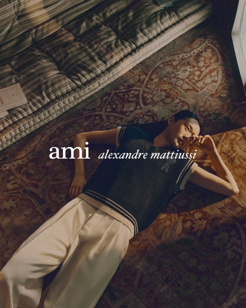 Model lying on patterned carpet wearing dark knit vest and light trousers for Ami Paris campaign