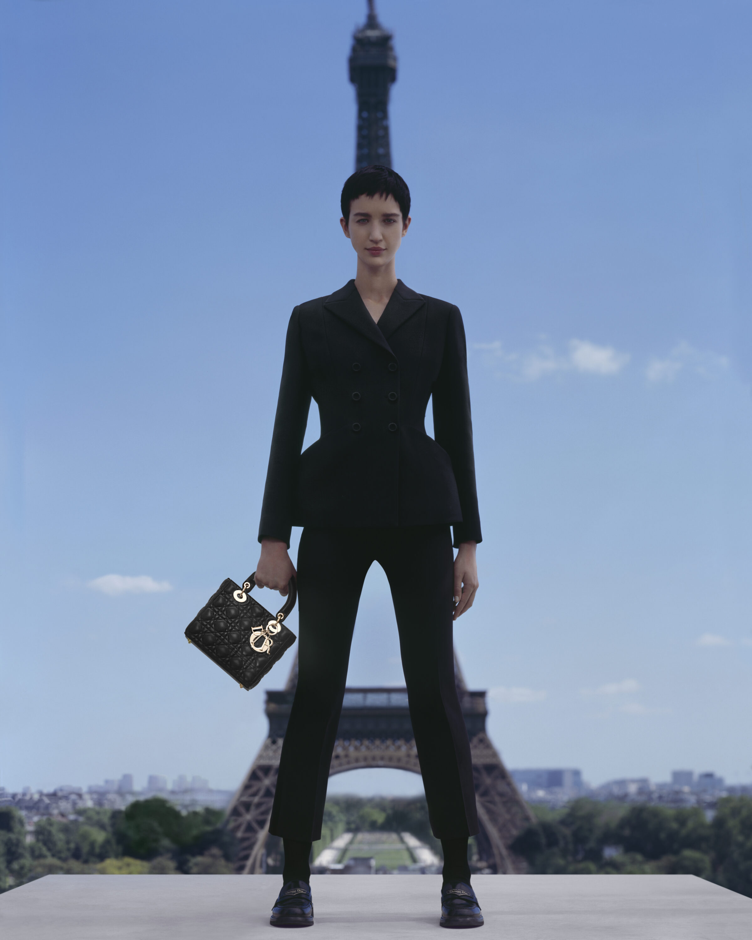Model in black suit holding a patterned handbag with the Eiffel Tower in the background